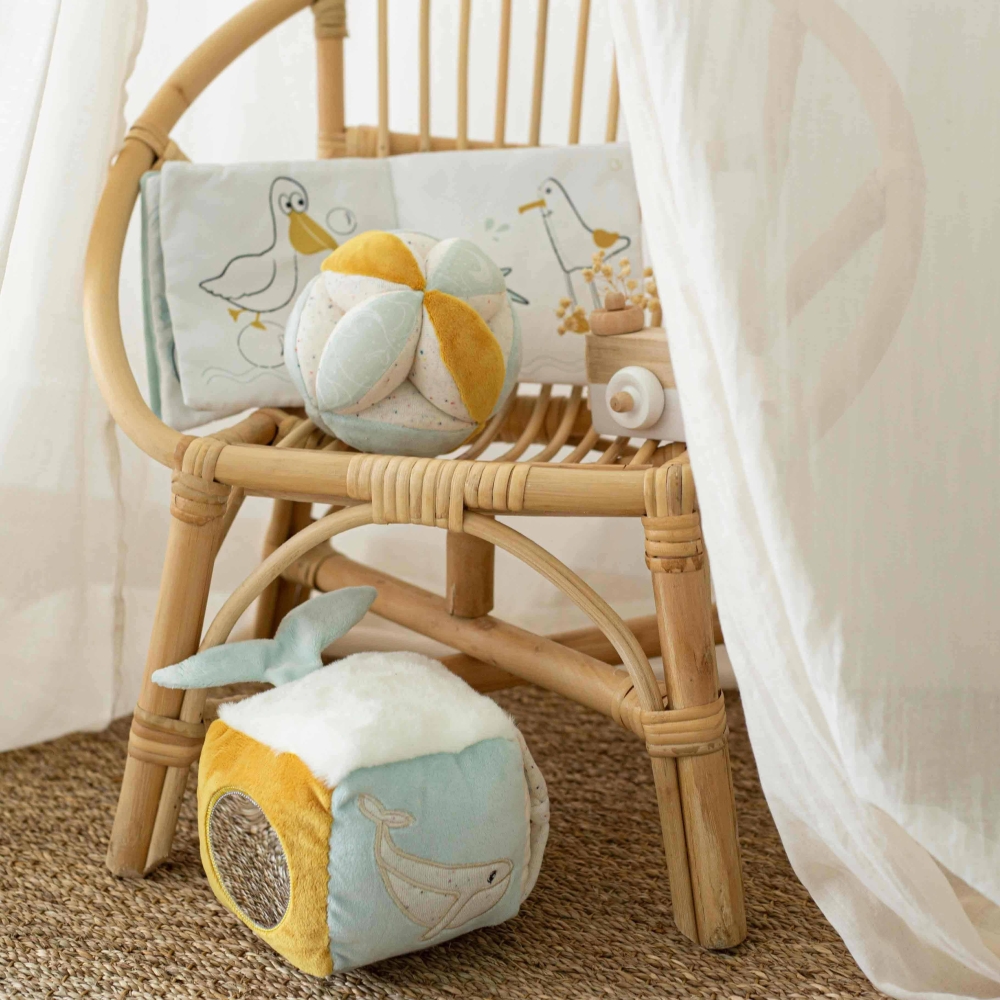 Cubo sensorial pousado no chão, com os outros brinquedos de estimulação da coleção numa cadeira de rotim Cubo sensorial pousado no chão, com os outros brinquedos de estimulação da coleção numa cadeira de rotim